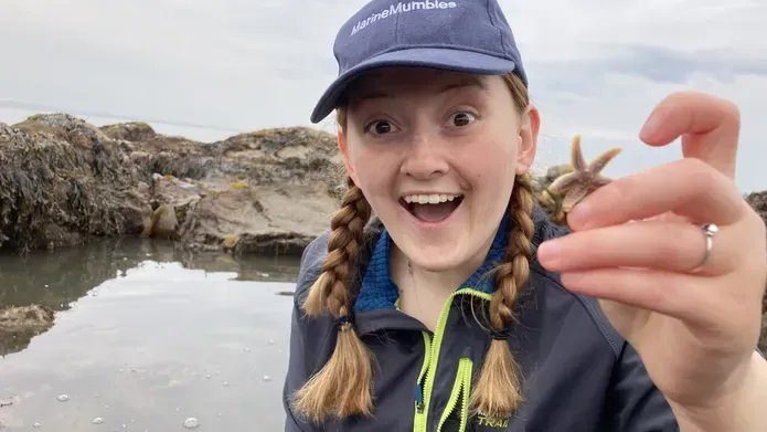 Close up of young woman in 20's sporting braided blonde hair, wearing a navy cap and jumper, excitedly holding up a small starfish against a background of water