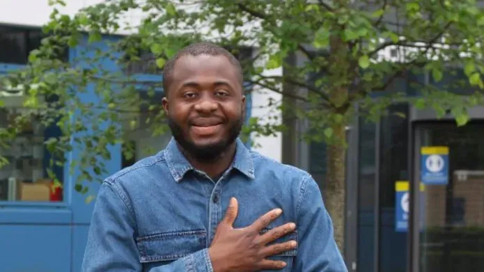 young black male in 20's wearing light blue shirt, posing with smile in front of green hedges and trees