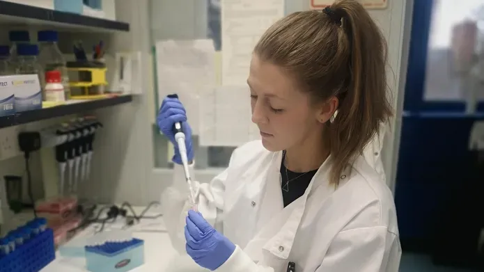 woman in lab coat and blue gloves holding pipette, background is a laboratory
