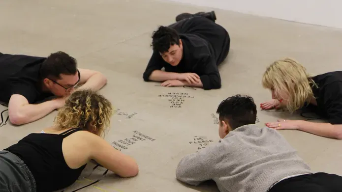 People lying face down on the floor in a circle looking at writing on the floor