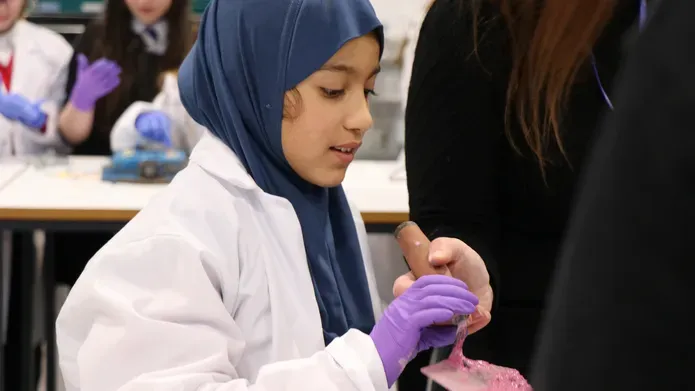 a young primary school girl wearing a navy headscarf and white lab coat, holding a shovel with mixed pink concrete