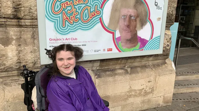 A young girl wearing a bright purple jacket, sat in a motored wheelchair, excitedly smiling in front of exhibition poster