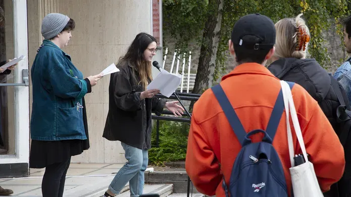 Audiences with Three white performers reading from scripts into a single microphone on the steps of the gallery underneath a decimal clock