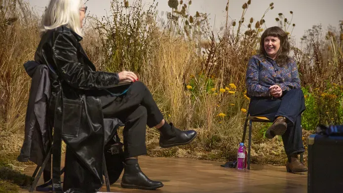 Two white woman talking physically distanced in a gallery backdropped by a meadow of dried flowers and grasses