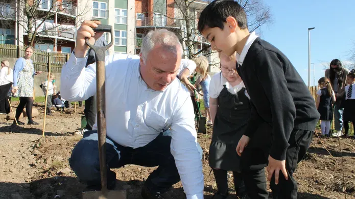 middle aged man crouched down on muddy land, balancing using a shovel, pointing to the ground while a young boy keenly looks on 