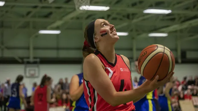 young woman in her 20's wearing red sporting tank top, holding orange basketball with one hand 