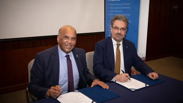two middle aged men, dressed formally in a suit and tie, sat beside one another at table both signing two pieces of white documents