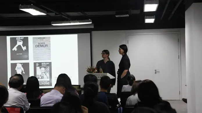 Two speakers stand at a podium in a room full of people.