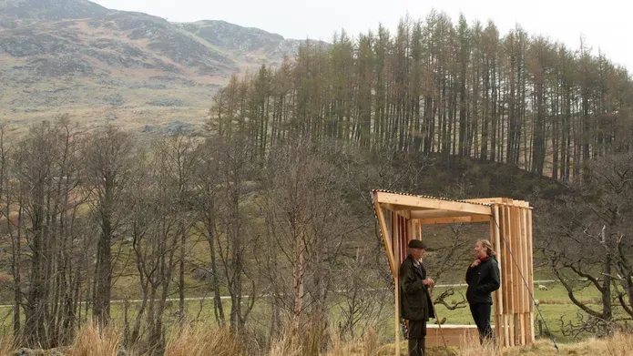 two people standing under a wooden structure in the middle of a grassy area with bare trees and rolling hills in background