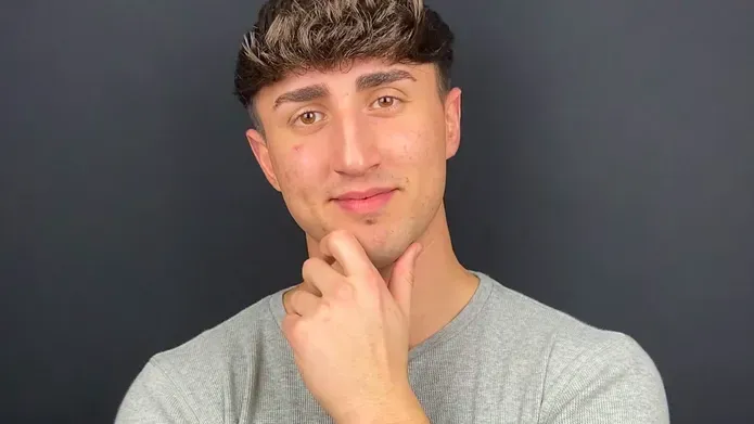 a young man with short dark hair holding his hand up to his chin while smiling, wearing a knitted grey jumper
