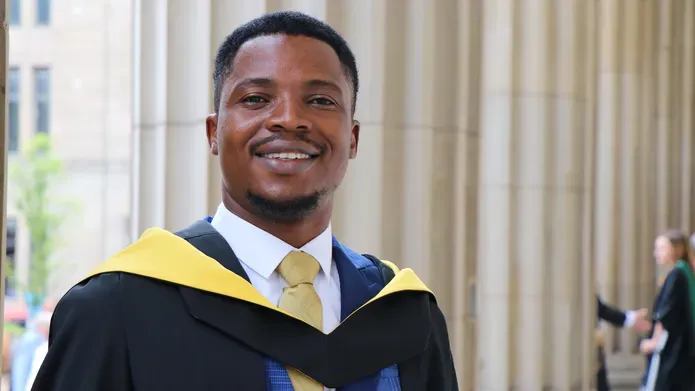young black man stands in graduation robes, posing against large stone pillars