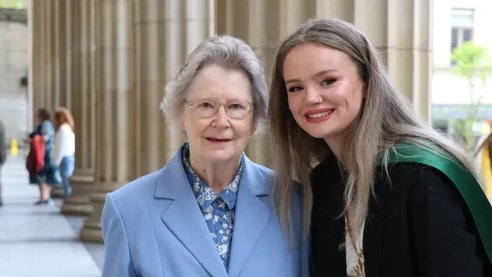 elderly woman wearing blue blazer standing next to young blonde woman wearing graduation grown