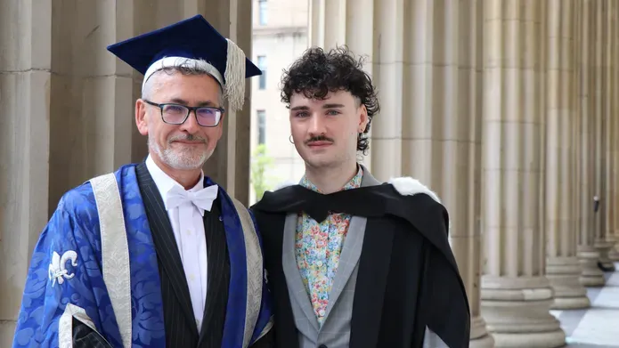 a middle aged man wearing a blue gown and tassled hat stands next to a young man in his 20's wearing a black graduation robe