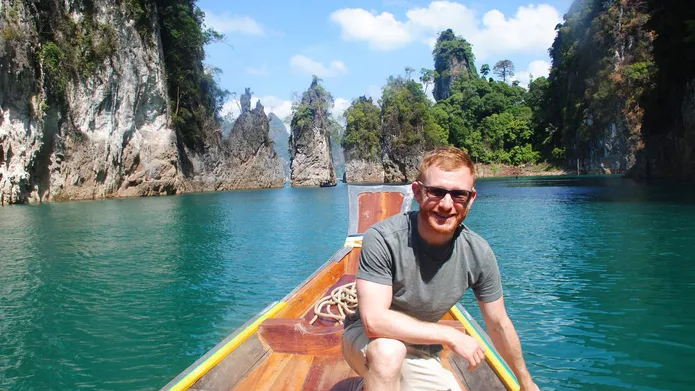 Ricardo Grammatica sits in a boat in a tropical location. Rocky cliffs and trees are in the background.