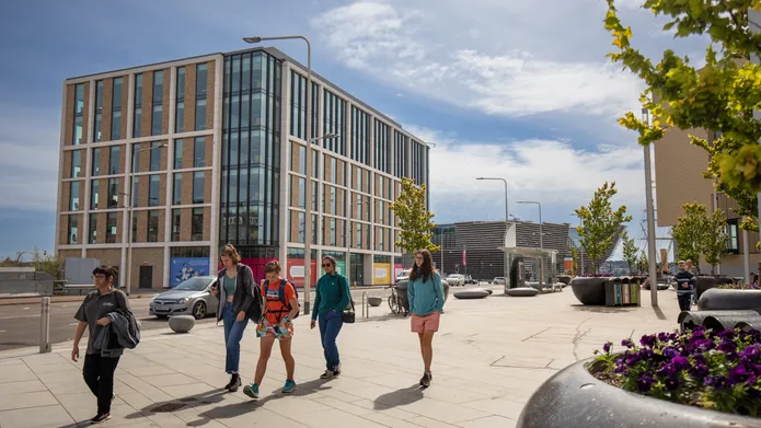A group of people walk in front of an office block on Dundee's waterfront with large trees in planters lining the pavement