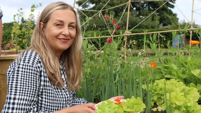 a woman with blonde hair, wearing a grey checkered shirt, holding a plate full of green salad beside a planter full of sprouting green plants 
