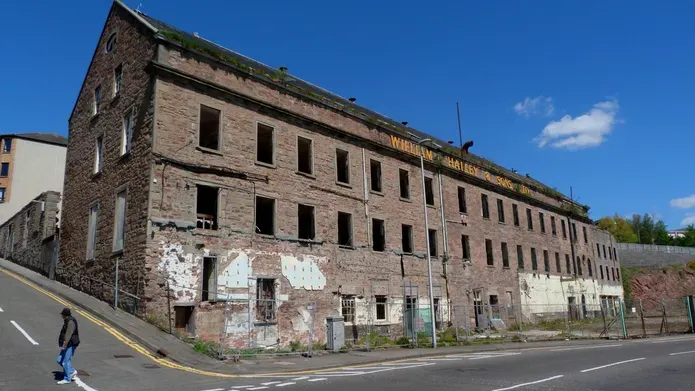 front view of a large derelict jute mill building on the edge of an empty road. The building is in a state of disrepair, with dark window openings on three different levels 