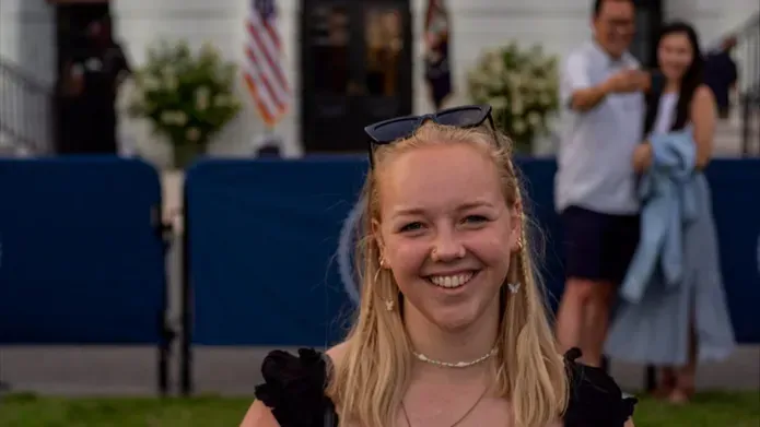 A young blonde girl in her early 20's, wearing a black short sleeved shirt, stands on grass in front of the White House. The American and Presidential Seal flags are in the background
