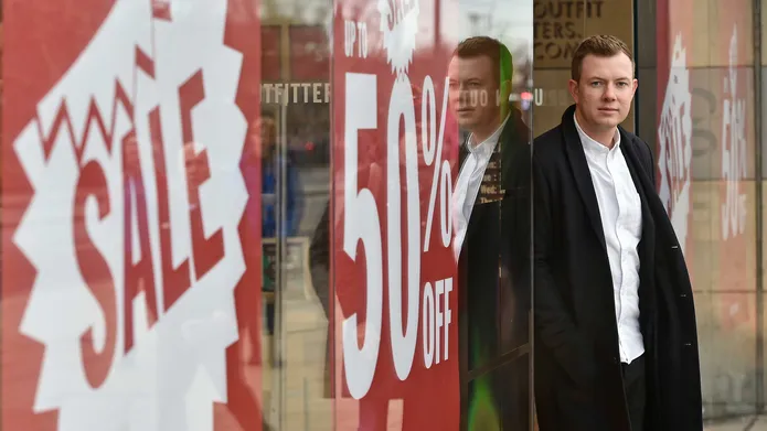 Cally Russell stands with his back to a shop window displaying red sale signs.