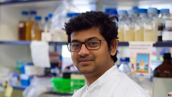 Joshua Peter standing in a lab with shelves full of bottles and lab equipment behind him