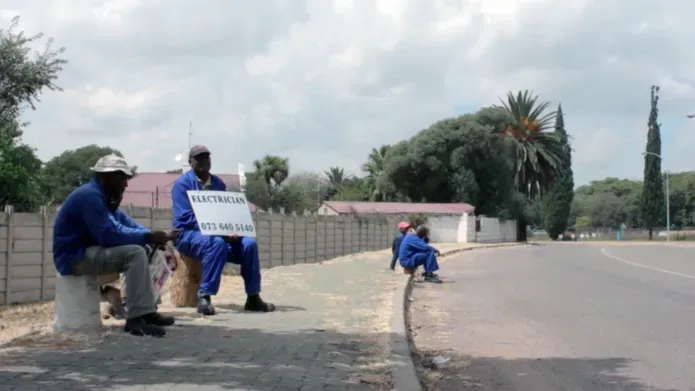 Black workers in blue overalls waiting in a road side with a sign reading electrician and a phone number