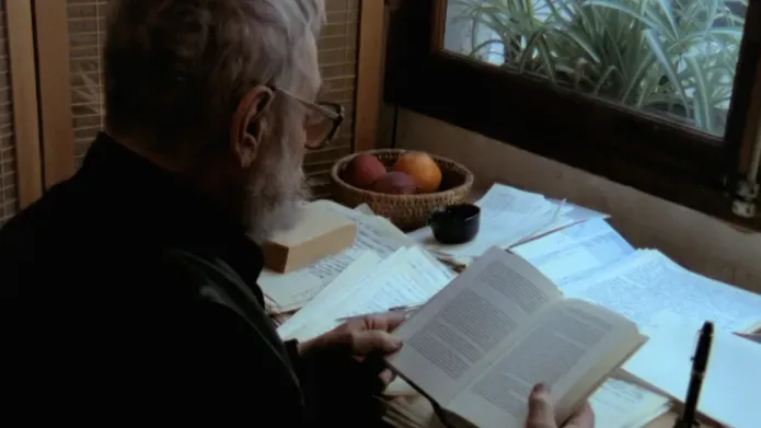 Film still. An elderly man with grey hair sits reading a book at a writing desk. 