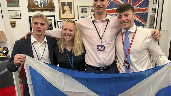 Four people stand together smiling, holding up the Scottish flag.