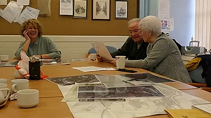 A female archivist and two older community members sit at a table discussing large photograph images of Dundee