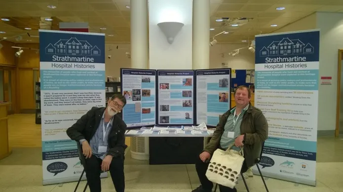 Two community members sit in front of a stall advertising the work of the Strathmartine Hospital Histories project.