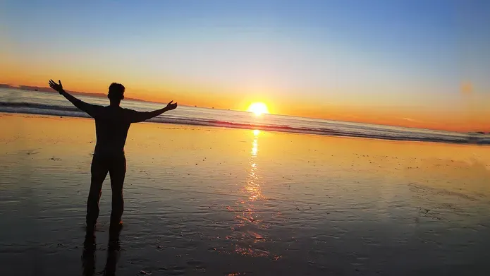 Person standing on beach at sunset with hands raised