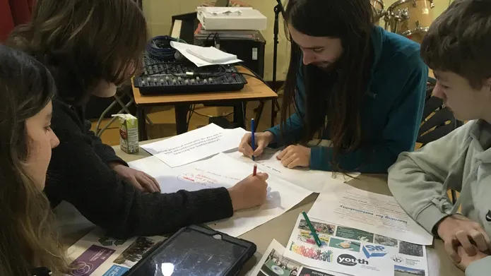 A group of four people sitting around a desk, working collaboratively, writing on pieces of paper.