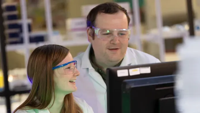 Two forensic scientists in lab equipment smiling as they view a computer screen.
