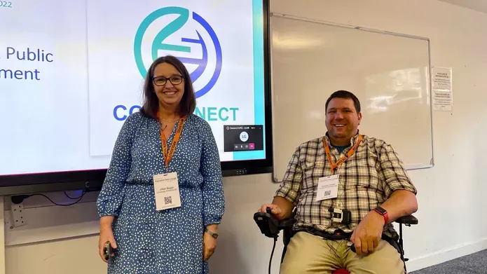 A woman in a blue dress stands next to a man in a wheel chair, in front of a presentation displayed on a screen.