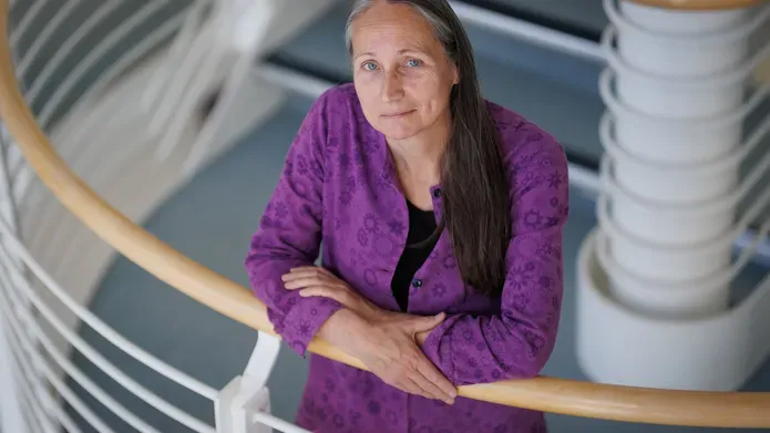 Kate Storey stands in a stairwell with her arms crossed leaning against the bannister, looking up.