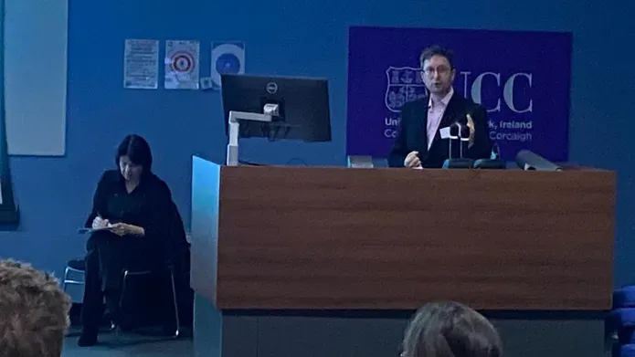 A man standing at a presentation stand, and a woman sits taking notes beside him.