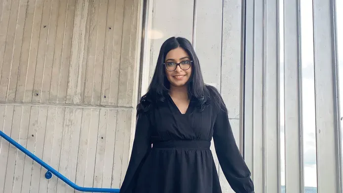 A woman standing in a stairwell by some windows, wearing a black dress.