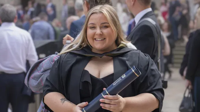 Smiling graduate with long hair, wearing gown and holding certificate tube