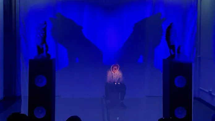 Heather Phillipson reading lit by the orange glow of her phone. She is bathed in blue light and flanked by two howling wolf sculptures and their looming shadows.