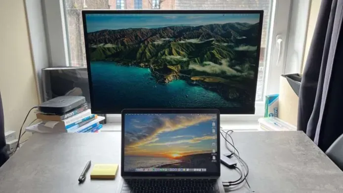 A laptop and computer monitor sit on a desk in front of a window. There is a pen and sticky notes sitting to the left on the desk.