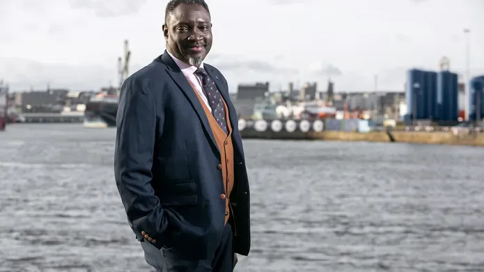 Ollie Folayan wearing a suit and waistcoat standing in front of a harbour.
