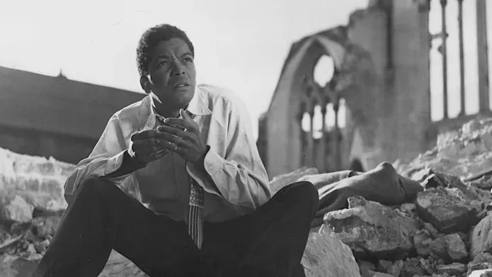 An actor sits on some rubble in front of the ruins of a church’s arched windows.