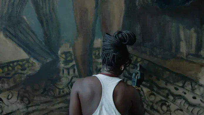 A black woman painting on a large canvas showing the legs of two people on a rug. The painter has a white vest and her braids are bunched in a bun.