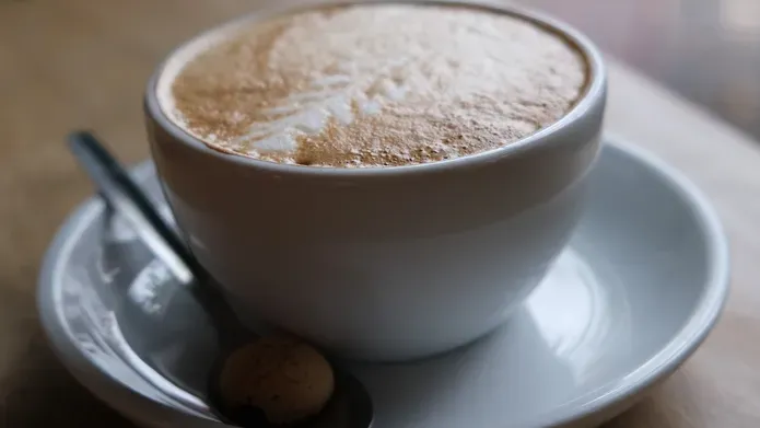 A photograph of a cup of coffee on a saucer with a teaspoon on a wooden table.