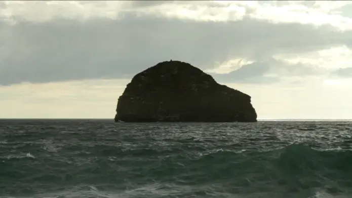 Film still. A rock in the ocean sits under a cloudy sky surrounded by a rough sea