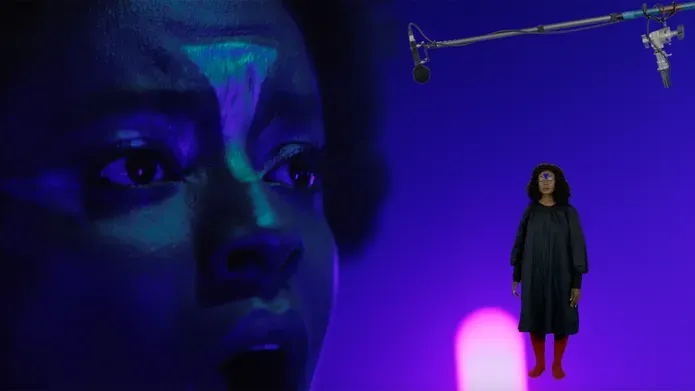 A close up of a black woman's face with a triangle painted on her forhead. Another woman with a triangle on her forehead is superimposed standing full height next to the looming head. A mic boom cuts across the image, the set is purple.
