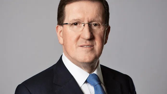 Man with brown hair and glasses wearing a dark suit with a white shirt with a blue tie is looking at the camera