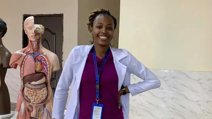 A woman wearing a purple dress and blazer, stands with her left hand on her hip. Anatomical models are behind her.