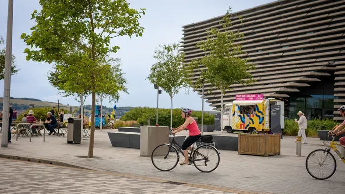 People sat a picnic tables outside V&A Dundee as cyclists go past