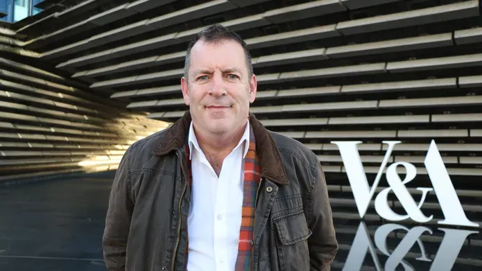 A man in a white shirt and brown jacket standing outside infront of the V&A museum