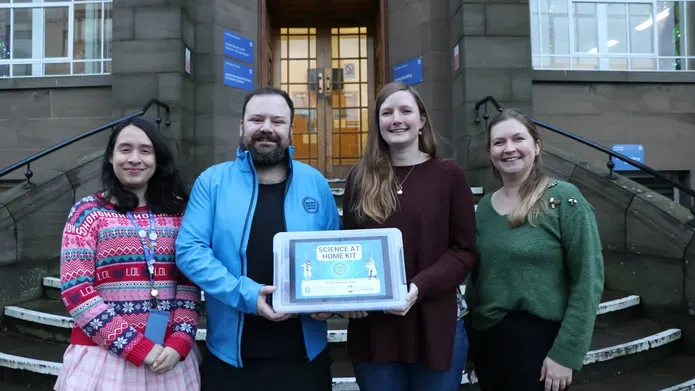 Four people stoof outside infront of steps, holding a plastic box with a blue and green label on it